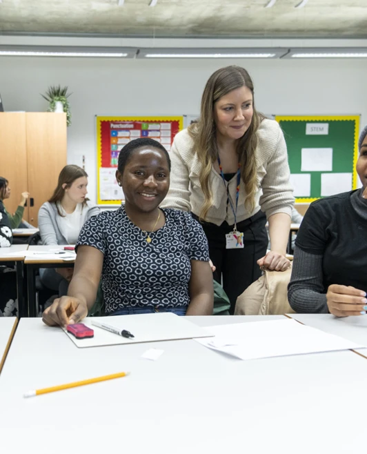A classroom scene where three female students are smiling and engaging in discussion. A teacher in a beige cardigan leans over the table to interact with them, while other students work in the background. A classroom scene where three female students are smiling and engaging in discussion. A teacher in a beige cardigan leans over the table to interact with them, while other students work in the background.